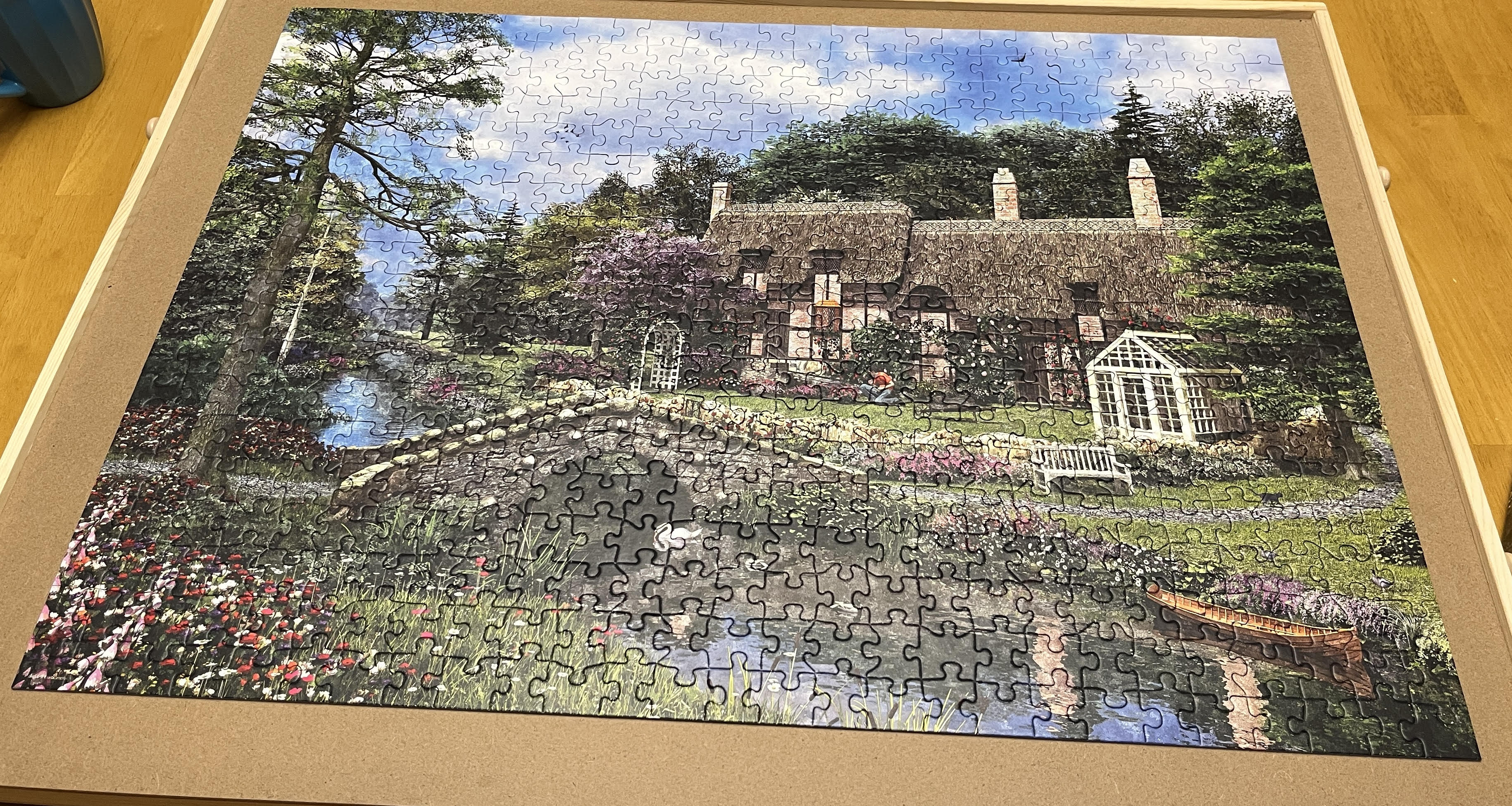 A picture of a completed puzzle on a puzzle board. The puzzle image is of a cottage in the English countryside, surrounded by flowers and a small brook with a bridge.